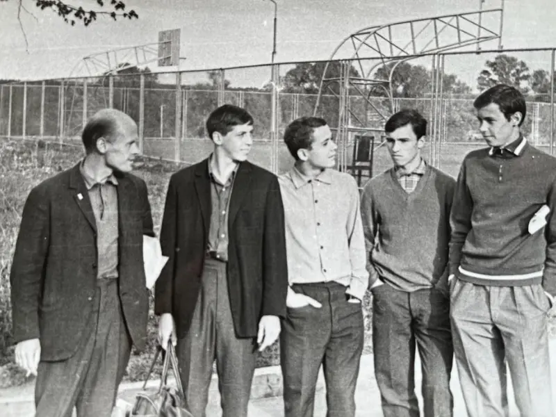 Grupa mężczyzn stoi przed ogrodzeniem, rozmawiając i spoglądając w kierunku obiektywu.