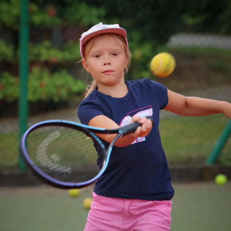 Młoda dziewczynka macha rakietą tenisową, przygotowując się do uderzenia piłki na korcie.