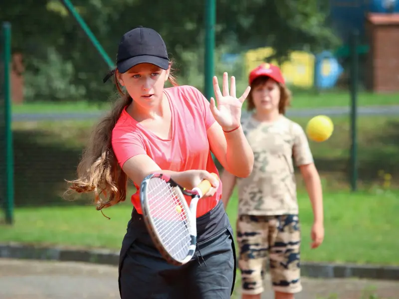 Młoda dziewczynka uderza rakietą tenisową w piłkę, pokazując energię i radość z gry na świeżym powietrzu.