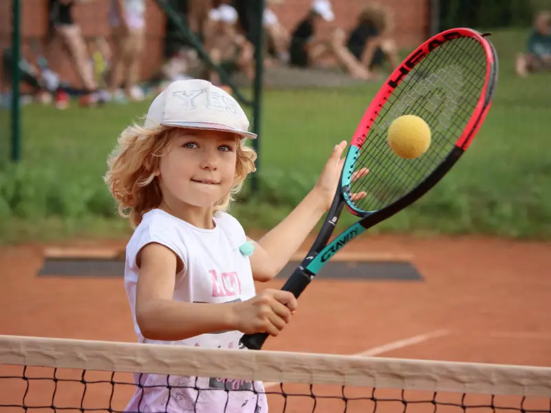 Młoda dziewczynka trzyma piłkę tenisową i rakietę tenisową, uśmiechając się z entuzjazmem na boisku.