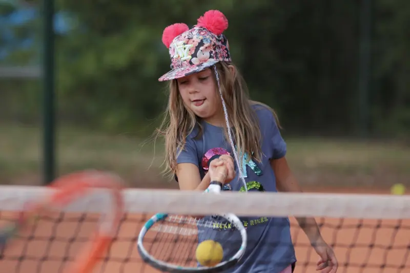 Młoda dziewczynka w kapeluszu grająca w tenisa na boisku, uśmiechnięta i pełna energii.