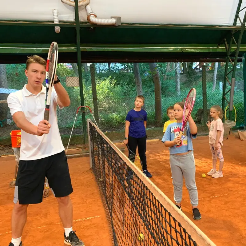 Mężczyzna trzymający rakietę tenisową przed grupą dzieci, które z zainteresowaniem obserwują jego ruchy.
