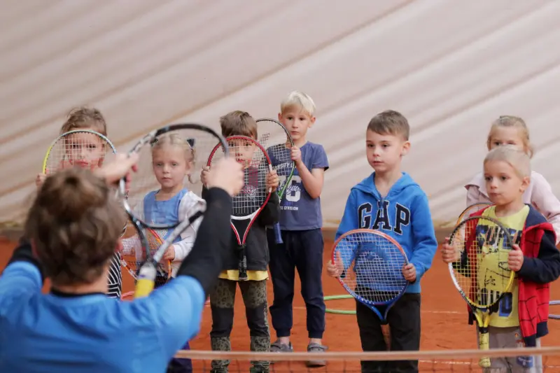 Grupa dzieci stoi wokół kortu tenisowego, obserwując grę i ciesząc się wspólnym czasem na świeżym powietrzu.