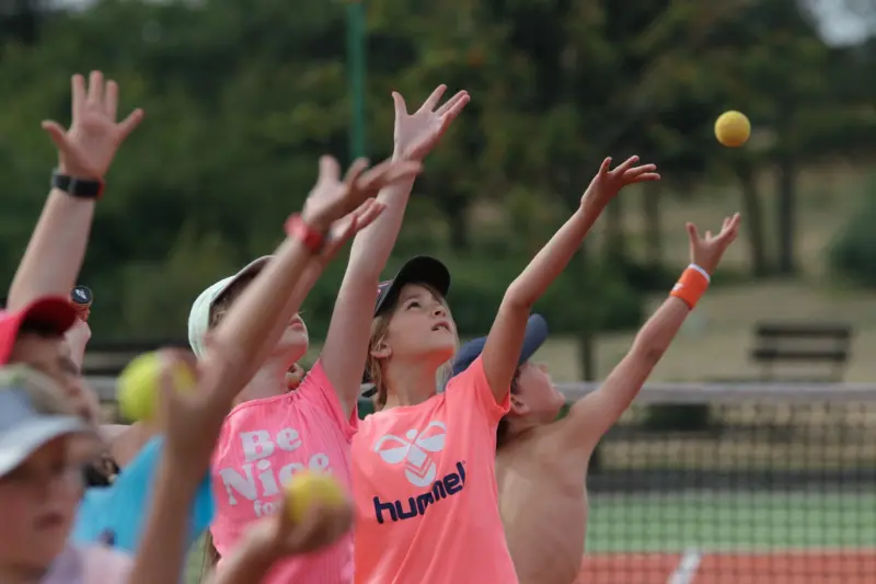 Grupa dzieci sięga w górę, próbując złapać tenisową piłkę w słonecznym otoczeniu.